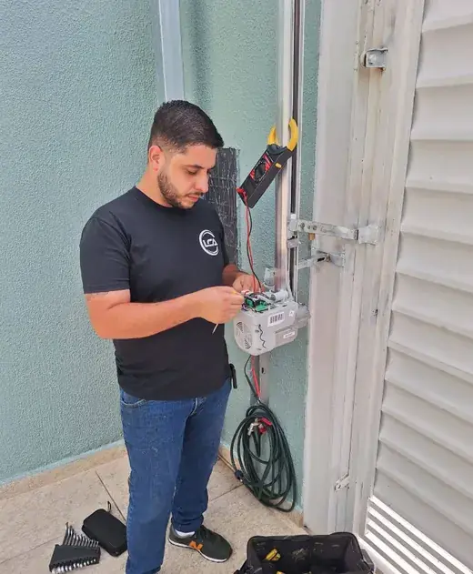 Técnico da LCA com camiseta da empresa ajustando a central de um motor de portão com alicate amperímetro